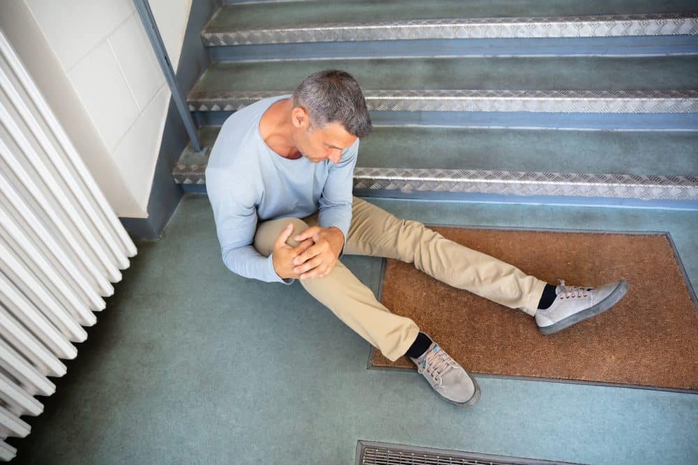 A man sitting at the bottom of a stairway holds his leg below the knee after a fall.
