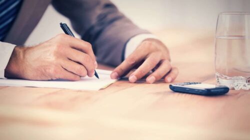 An attorney’s hands filling out paperwork. A calculator and glass of water sit on the table nearby.