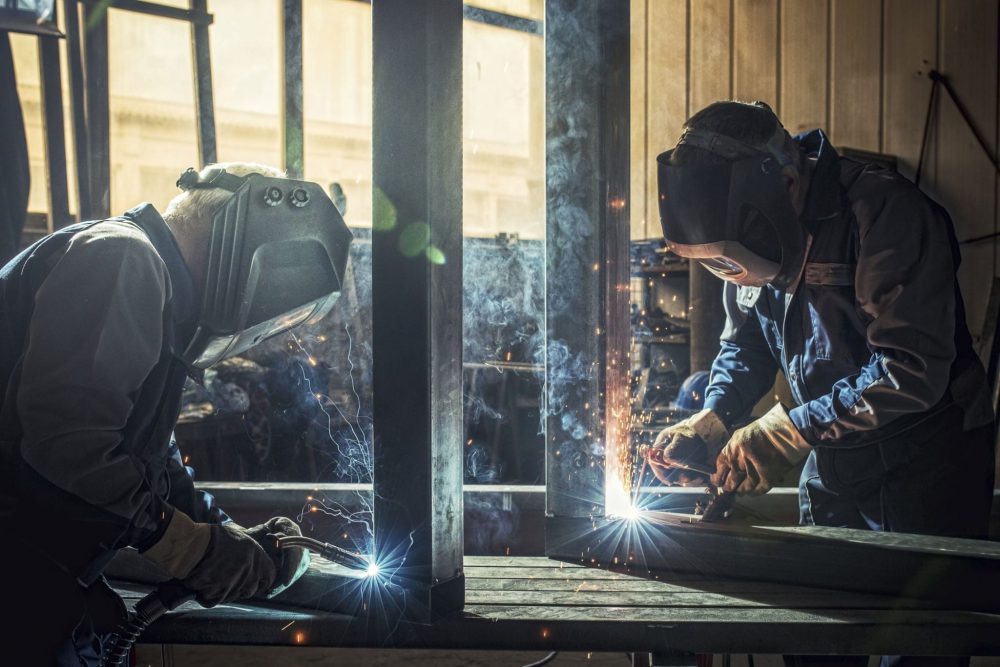 Two welders wearing protective helmets and gloves are working with metal, using welding torches in a workshop setting.