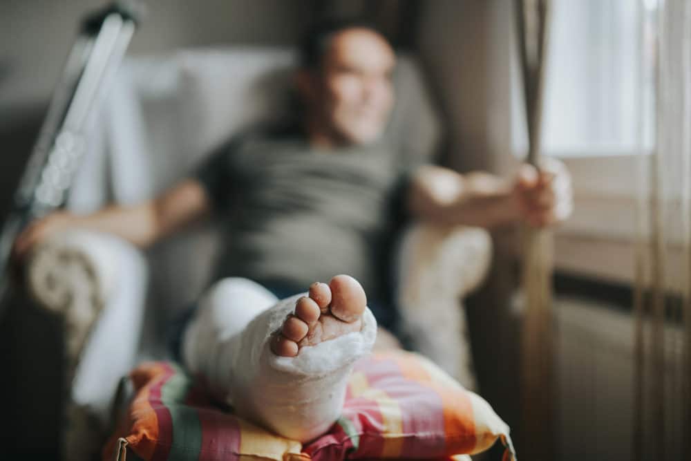 A man sits in a chair with his leg elevated and wrapped in a cast, crutches leaning against the chair. The focus is on the injured leg in the foreground.