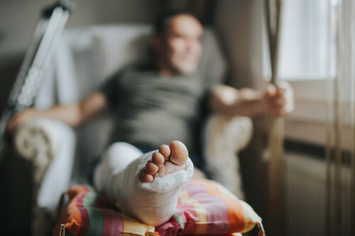 A man sits in a chair with his leg elevated and wrapped in a cast, crutches leaning against the chair. The focus is on the injured leg in the foreground.